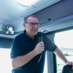 Jim Lollar stands at the front of a bus holding a microphone