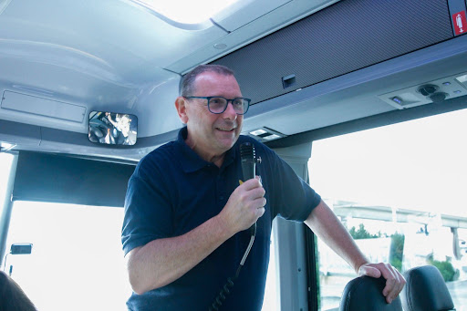 Jim Lollar stands at the front of a bus holding a microphone